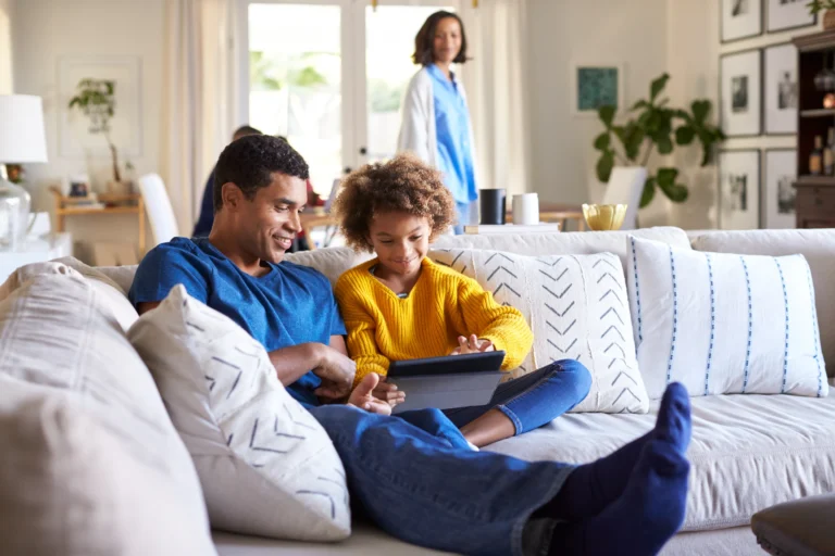 A man and a child sit on a sofa using a tablet, while another person stands in the background discussing heat pump maintenance. The room is well-lit and decorated with plants and wall art, creating a cozy atmosphere.