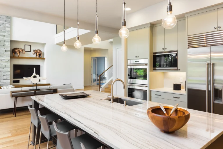 Modern kitchen with marble island, bar stools, and pendant lights. Sleek kitchen plumbing complements stainless steel appliances and a wooden bowl on the counter. Open layout to a living area with tasteful decor and large windows.