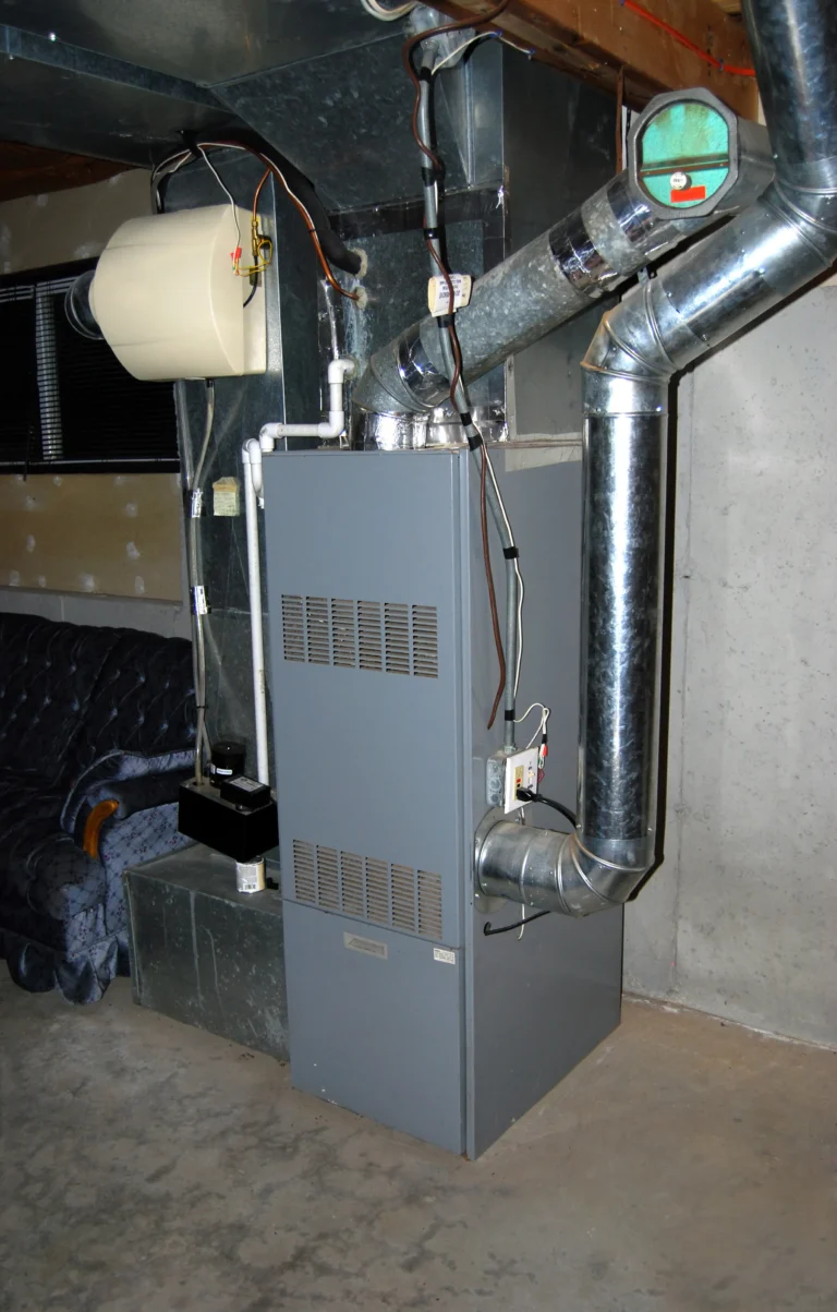 A residential basement showcases a recently completed furnace install, featuring a gray furnace and ductwork system, with a dehumidifier unit mounted on top. These functional elements sit beside a comfortable blue couch, creating both an efficient and inviting space.