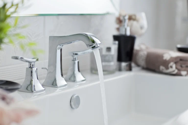 Modern bathroom sink with chrome faucet and handles, water running gracefully. Hints of expert plumbing ensure efficiency, while blurred toiletries in the background and neatly folded towels to the side add a touch of elegance.